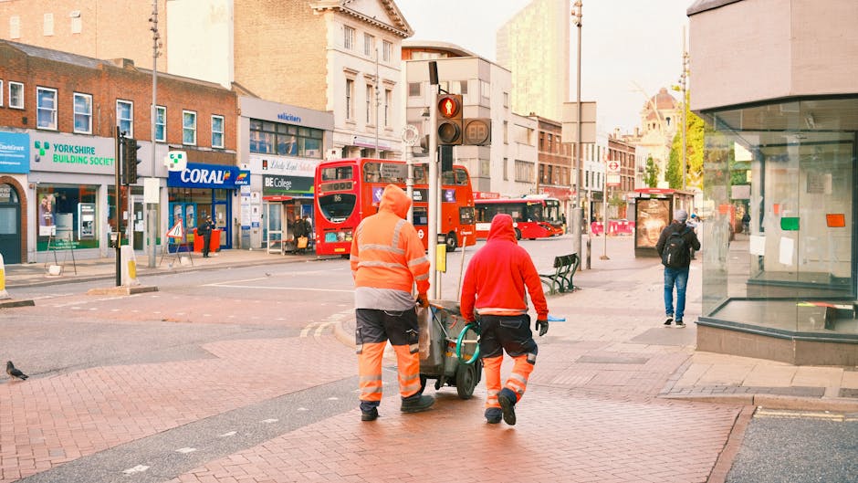 A busy urban scene near Bromley South station featuring two maintenance workers in high-visibility orange and red jackets and trousers, pushing a street cleaning cart along a paved sidewalk. The backg