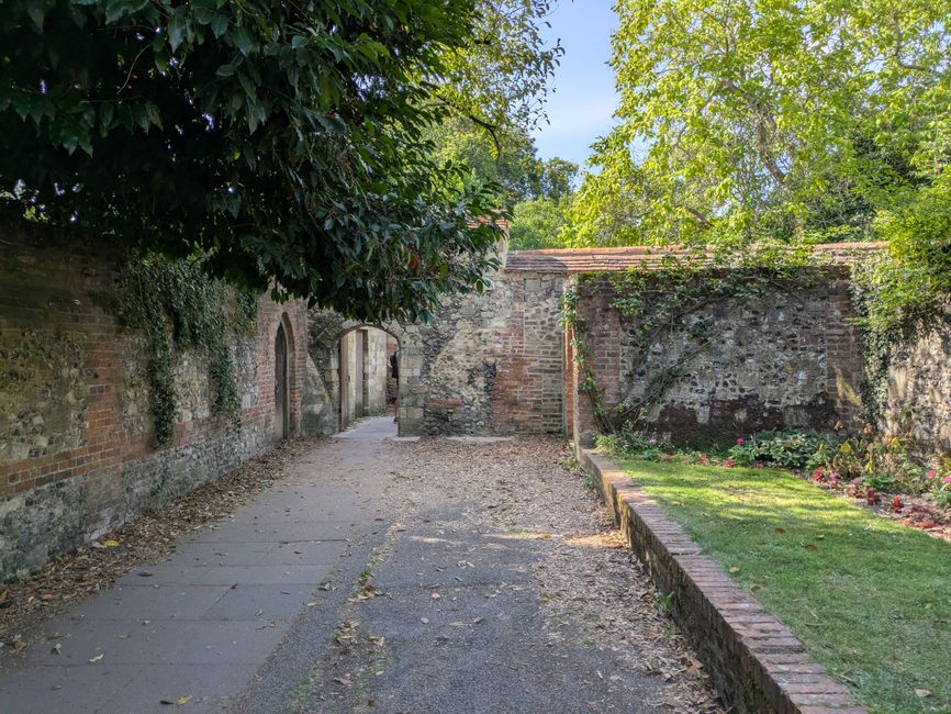 A narrow garden pathway paved with uneven bricks, winding through a lush, well-maintained garden enclosed by brick pillars and curved brick walls. The pathway is bordered by dense green shrubs, includ