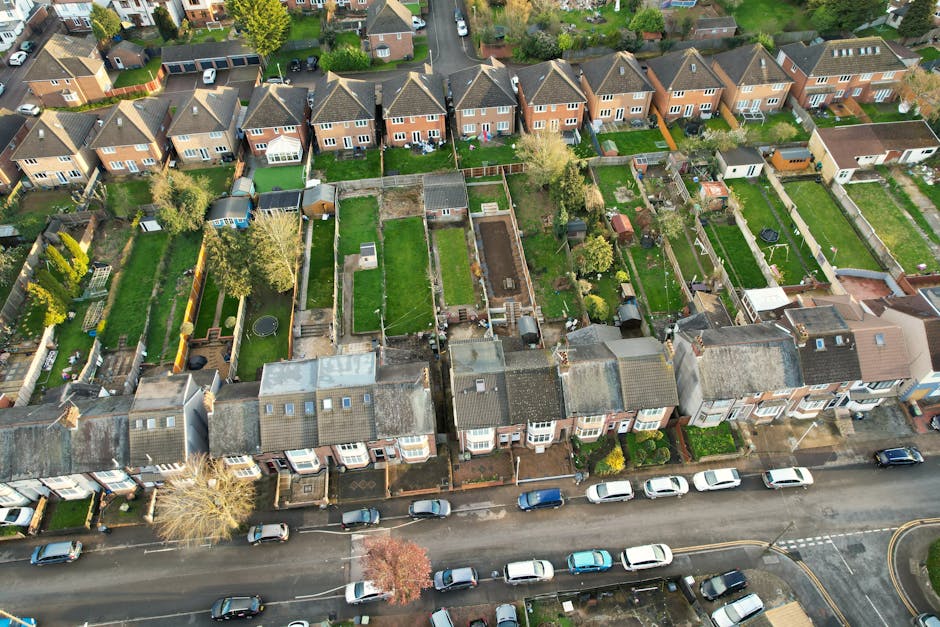 An aerial view of a row of terraced homes with small, well-maintained gardens at the rear. Each garden features different layouts, including lush green lawns, flower beds, paved pathways, and vegetabl